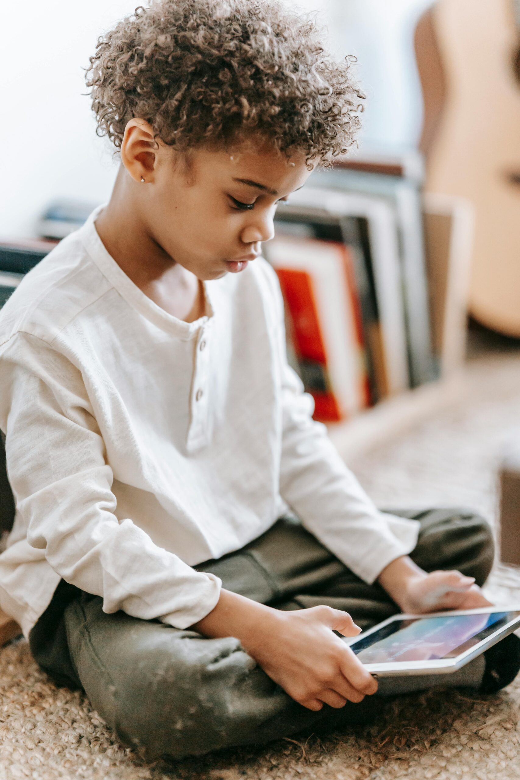 Full body side view of concentrated Africa American boy playing video game on tablet while sitting with crossed legs near books in room with guitar on blurred background
